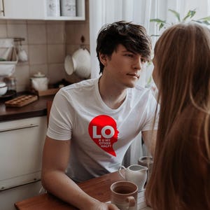 Puede incluir: Una camiseta blanca de manga corta con un gr&aacute;fico rojo en forma de coraz&oacute;n en el pecho. El gr&aacute;fico contiene el texto blanco LO y el texto m&aacute;s peque&ntilde;o R U MY OTHER HALF? dentro del coraz&oacute;n, que forma una silueta de un rostro.