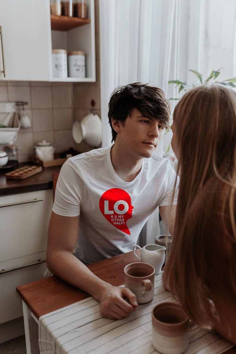 Puede incluir: Una camiseta blanca de manga corta con un gr&aacute;fico rojo en forma de coraz&oacute;n en el pecho. El gr&aacute;fico contiene el texto blanco LO y el texto m&aacute;s peque&ntilde;o R U MY OTHER HALF? dentro del coraz&oacute;n, que forma una silueta de un rostro.