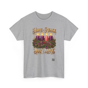 May include: A heather gray t-shirt featuring a graphic of four lit candles nestled in a green wreath with purple ribbons. The text above reads HOPE PEACE and the text below reads JOY LOVE in a gold font. A small QR code is visible at the bottom.