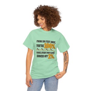 May include: A light green short-sleeve t-shirt featuring black and yellow text that reads: FROM 100 FEET AWAY, YOU'RE 100%. SADLY, EVERY FOOT CLOSER KNOCKS OFF 1%. The design includes a graphic of small footprints along a horizontal line.