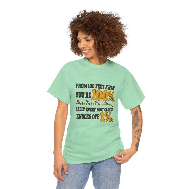 May include: A light green short-sleeve t-shirt featuring black and yellow text that reads: FROM 100 FEET AWAY, YOU'RE 100%. SADLY, EVERY FOOT CLOSER KNOCKS OFF 1%. The design includes a graphic of small footprints along a horizontal line.