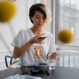 May include: A white short-sleeved t-shirt featuring a graphic of an orange tabby cat sitting in a lotus position with the text "LESS DRAMA, MORE KARMA" printed above and below the cat illustration in a brown, stylized font.
