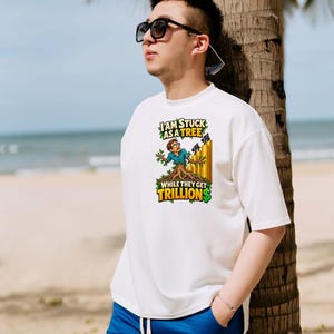 May include: A white short-sleeved t-shirt featuring a graphic of a person transformed into a tree with roots, standing next to a stack of gold coins. The text reads, I AM STUCK AS A TREE WHILE THEY GET TRILLIONS, with a dollar sign in green.