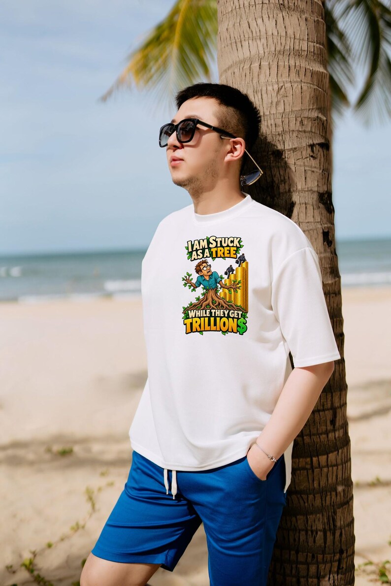 May include: A white short-sleeved t-shirt featuring a graphic of a person transformed into a tree with roots, standing next to a stack of gold coins. The text reads, I AM STUCK AS A TREE WHILE THEY GET TRILLIONS, with a dollar sign in green.