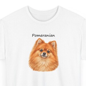 May include: A white t-shirt featuring a detailed illustration of a fluffy orange Pomeranian dog centered on the chest. Above the dog's head, the word Pomeranian is printed in a simple, black, sans-serif font. The shirt has a crew neckline.