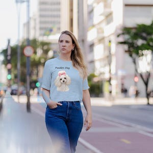May include: A light blue short-sleeved t-shirt featuring a graphic of a white poodle with a pink bow on its head and the text Poodle's Day printed above the dog illustration in a simple black font.