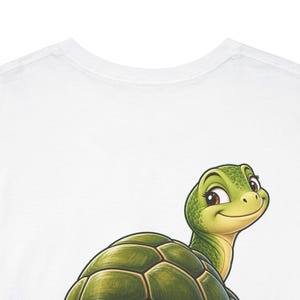May include: A white t-shirt featuring a graphic illustration of a smiling green turtle with a textured shell. The turtle is positioned in the lower portion of the shirt, looking upward with large dark eyes and a friendly expression.