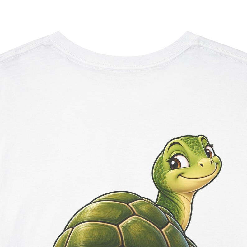 May include: A white t-shirt featuring a graphic illustration of a smiling green turtle with a textured shell. The turtle is positioned in the lower portion of the shirt, looking upward with large dark eyes and a friendly expression.