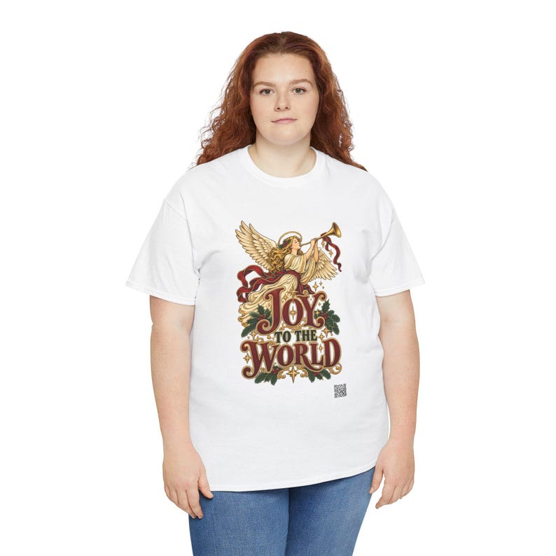 May include: A white short-sleeve t-shirt featuring a graphic of an angel playing a trumpet with the text Joy to the World in red and gold lettering, surrounded by green holly leaves and red berries, with a small QR code at the bottom.