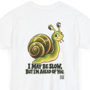 May include: A white t-shirt featuring a cartoon snail with a green body and a dark green spiral shell. Below the snail, the text reads, "I MAY BE SLOW, BUT I'M AHEAD OF YOU." A small QR code is printed in the bottom right corner of the graphic.