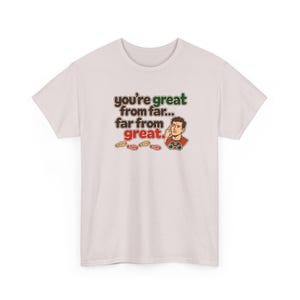 May include: A light pink t-shirt featuring a graphic of a person looking through binoculars with the text "you're great from far... far from great." and small footprints below the illustration in brown, green, and red ink.
