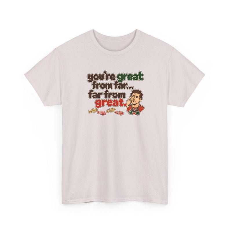 May include: A light pink t-shirt featuring a graphic of a person looking through binoculars with the text "you're great from far... far from great." and small footprints below the illustration in brown, green, and red ink.