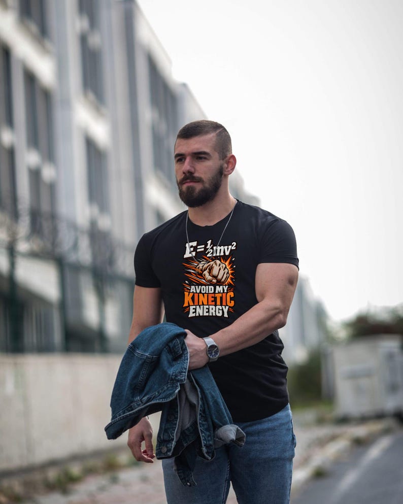 Puede incluir: Una camiseta negra de manga corta con un estampado gr&aacute;fico de un pu&ntilde;o con efecto de explosi&oacute;n, la ecuaci&oacute;n f&iacute;sica E=1/2mv&sup2; y el texto AVOID MY KINETIC ENERGY en letras blancas y naranjas en el pecho.