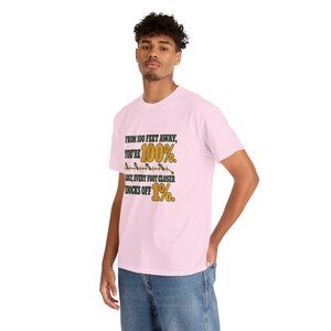 May include: A light pink short-sleeve t-shirt featuring the black and gold text: FROM 100 FEET AWAY, YOU'RE 100%. SADLY, EVERY FOOT CLOSER KNOCKS OFF 1%. The design includes a small graphic of a measuring line with footprints.