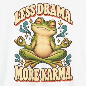 May include: A white t-shirt featuring a green frog sitting in a lotus position with eyes closed. The text "LESS DRAMA." is arched above the frog, and "MORE KARMA." is below. The design includes small yellow stars and teal swirls.