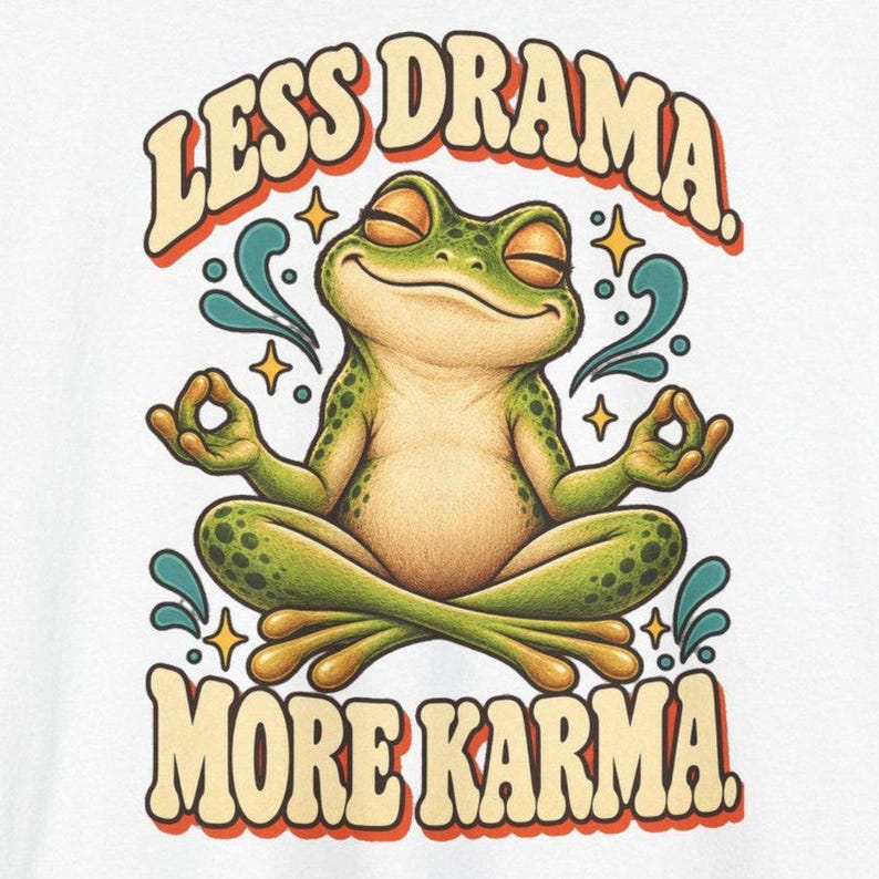 May include: A white t-shirt featuring a green frog sitting in a lotus position with eyes closed. The text "LESS DRAMA." is arched above the frog, and "MORE KARMA." is below. The design includes small yellow stars and teal swirls.