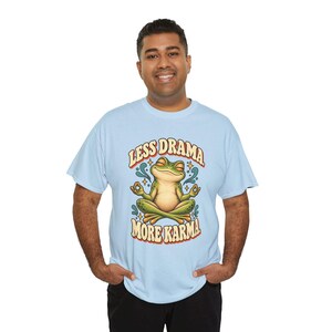 May include: A light blue short-sleeve t-shirt featuring a graphic of a green frog sitting in a lotus position with eyes closed. The text above the frog reads LESS DRAMA and the text below reads MORE KARMA in a bold, retro-style font.