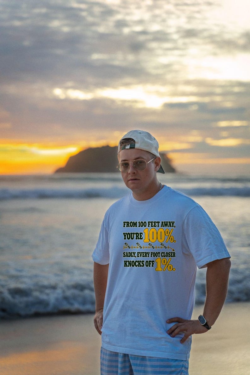 May include: A white short-sleeved t-shirt featuring the text "FROM 100 FEET AWAY, YOU'RE 100%. SADLY, EVERY FOOT CLOSER KNOCKS OFF 1%." The text is printed in black and yellow, with a small graphic of a measuring line between the lines of text.
