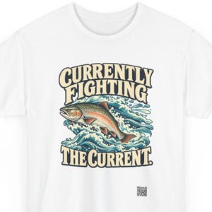 May include: A white t-shirt featuring a graphic of a trout jumping through blue and white stylized waves. The text above the fish reads CURRENTLY FIGHTING and the text below reads THE CURRENT. A small QR code is printed near the bottom right.