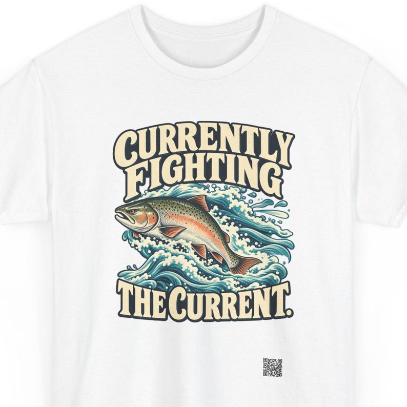 May include: A white t-shirt featuring a graphic of a trout jumping through blue and white stylized waves. The text above the fish reads CURRENTLY FIGHTING and the text below reads THE CURRENT. A small QR code is printed near the bottom right.