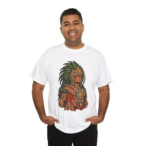 May include: A white short-sleeved t-shirt featuring a detailed graphic print of an Aztec warrior wearing an elaborate green feathered headdress, a sun-disk chest piece, and traditional red and green patterned garments on the front center.