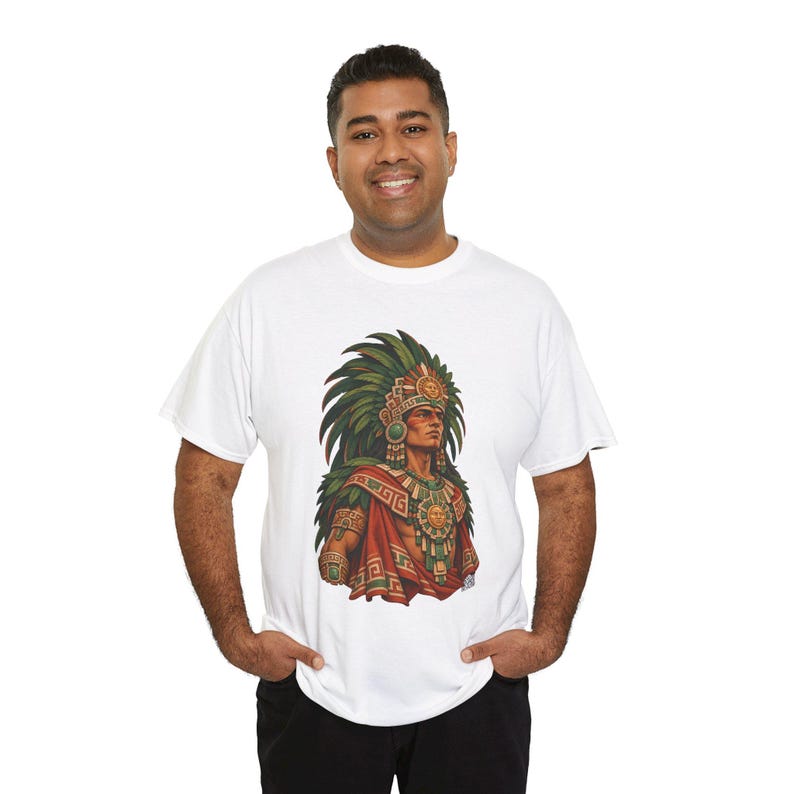 May include: A white short-sleeved t-shirt featuring a detailed graphic print of an Aztec warrior wearing an elaborate green feathered headdress, a sun-disk chest piece, and traditional red and green patterned garments on the front center.