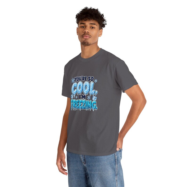 May include: A dark gray short-sleeve t-shirt featuring a graphic print with the text "YOU'RE SO COOL, I'M FREEZING." The lettering is styled in a light blue icy font, surrounded by small white snowflake icons and ice cube illustrations.