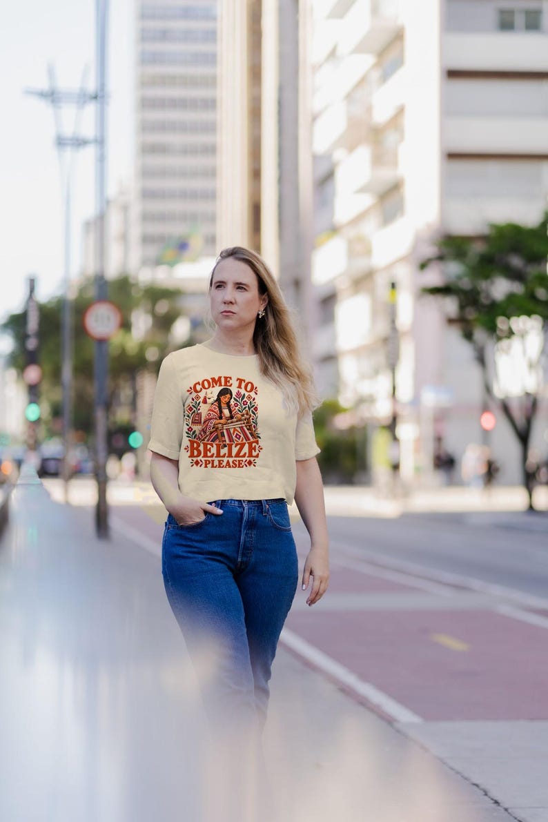 May include: A cream-colored short-sleeve t-shirt featuring a graphic illustration of a person weaving, surrounded by colorful floral patterns and the text "COME TO BELIZE PLEASE" printed in red and black lettering on the front chest.