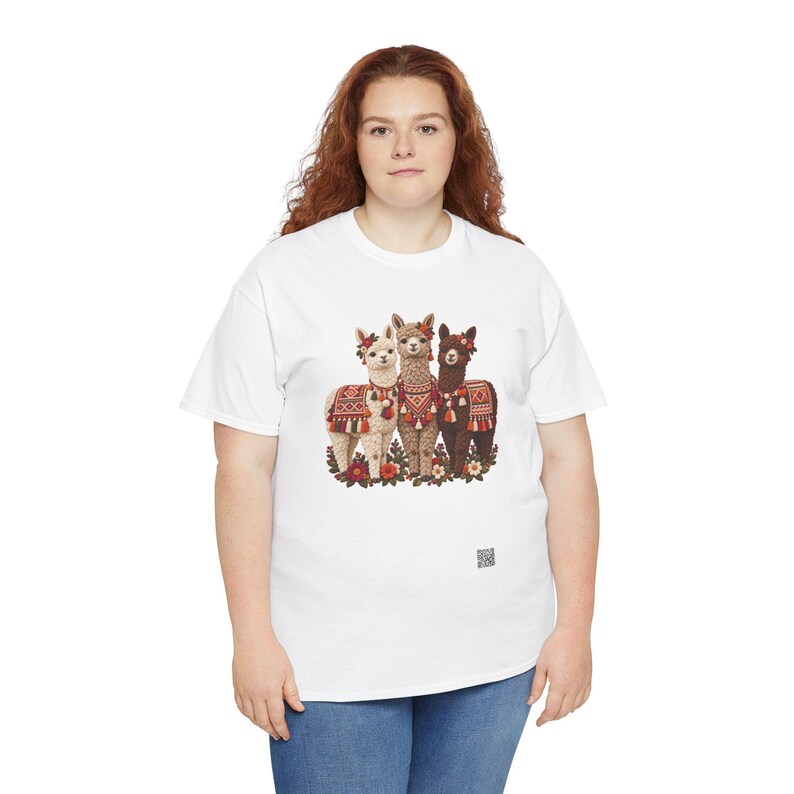 May include: A white t-shirt featuring a graphic print of three alpacas standing together. The alpacas are adorned with colorful patterned blankets, tassels, and floral headpieces. The design includes red, brown, and cream tones.