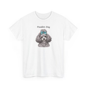Puede incluir: Una camiseta blanca de manga corta con el gr&aacute;fico de un caniche gris con un lazo azul brillante en la cabeza. Sobre el caniche, el texto Poodle's Day est&aacute; impreso en una fuente negra sencilla. La camiseta tiene cuello redondo y corte est&aacute;ndar.