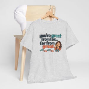 May include: A light gray t-shirt featuring a graphic illustration of a person with binoculars and the text "you're great from far... far from great." The design includes colorful footprints in teal and orange, paired with a plain white baseball cap.