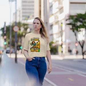 May include: A beige short-sleeved t-shirt featuring a graphic of a person integrated into a tree trunk. The text reads "I AM STUCK AS A TREE" and "WHILE THEY GET TRILLION$" in green and yellow lettering with a fence background.