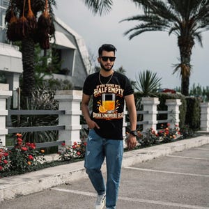 May include: A black short-sleeve t-shirt featuring a graphic of a glass of orange juice with an orange slice and the text IF YOU THINK IT'S HALF EMPTY DRINK SOMEWHERE ELSE, paired with blue denim jeans and white athletic sneakers.