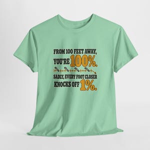 May include: A light green short-sleeve t-shirt featuring the black and orange text: FROM 100 FEET AWAY, YOU'RE 100%. SADLY, EVERY FOOT CLOSER KNOCKS OFF 1%. The design includes a horizontal line with small footprint icons and a downward arrow.