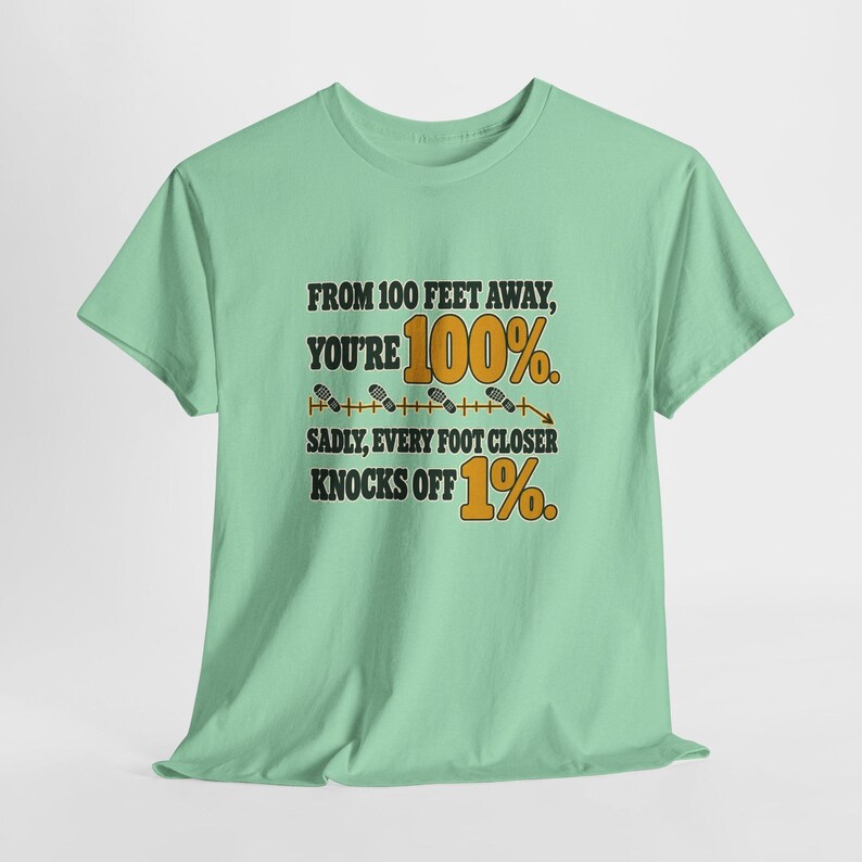 May include: A light green short-sleeve t-shirt featuring the black and orange text: FROM 100 FEET AWAY, YOU'RE 100%. SADLY, EVERY FOOT CLOSER KNOCKS OFF 1%. The design includes a horizontal line with small footprint icons and a downward arrow.