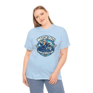 May include: A light blue t-shirt featuring a graphic of a pilot with headphones and a small robot in a cockpit. The text reads WATCH OUT! I'M ON AUTOPILOT NOW. The design is circular with wings and a small airplane icon at the bottom.
