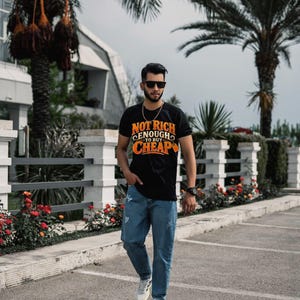May include: A black short-sleeve t-shirt featuring the orange and white text "NOT RICH ENOUGH TO BUY CHEAP" on the chest, paired with blue distressed denim jeans and white sneakers. The shirt has a crew neckline and a graphic print design.