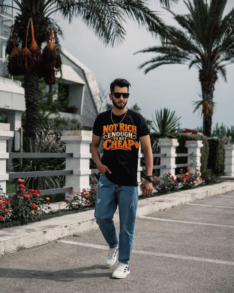 May include: A black short-sleeve t-shirt featuring the orange and white text "NOT RICH ENOUGH TO BUY CHEAP" on the chest, paired with blue distressed denim jeans and white sneakers. The shirt has a crew neckline and a graphic print design.