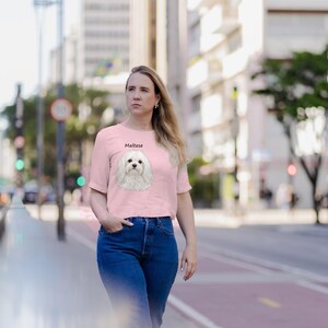 May include: A light pink short-sleeved t-shirt featuring a graphic illustration of a white Maltese dog centered on the chest with the word Maltese printed in black text above the dog's head.