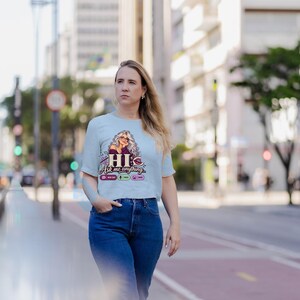 May include: A light blue short-sleeved t-shirt featuring a graphic illustration of a woman with long wavy hair and glasses. The text reads HI Ask me anything, accompanied by small icons for new chat, voice, and image functions.