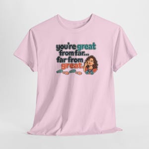 May include: A light pink short-sleeve t-shirt featuring a graphic print with the text "you're great from far... far from great." The design includes a cartoon illustration of a person with binoculars and several small colorful footprints.