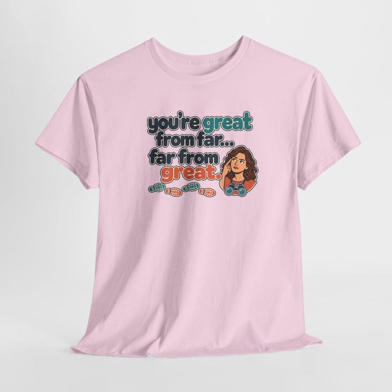 May include: A light pink short-sleeve t-shirt featuring a graphic print with the text "you're great from far... far from great." The design includes a cartoon illustration of a person with binoculars and several small colorful footprints.