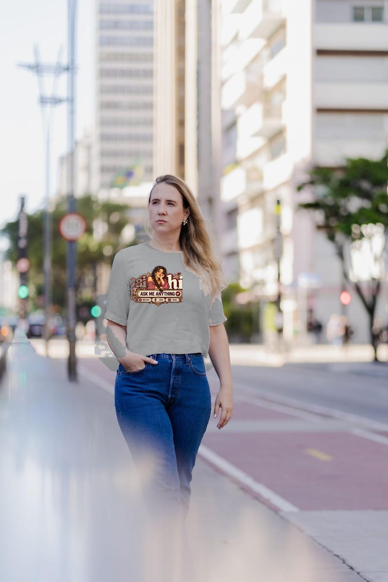 May include: A light gray short-sleeved t-shirt featuring a graphic print of a person with long dark hair, the text "hi" in a stylized font, and a rectangular box containing the text "ASK ME ANYTHING" with small circular icons.