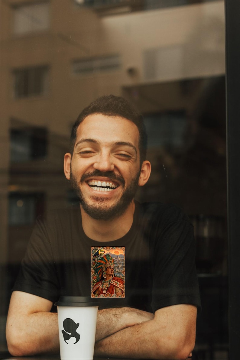 May include: A black t-shirt featuring a rectangular graphic print of a person wearing an elaborate feathered headdress and traditional attire, with a toucan perched nearby against a sunset background, positioned above a white coffee cup.