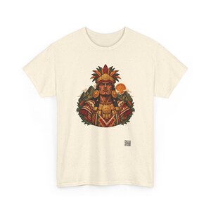May include: A cream-colored t-shirt featuring a central graphic of a person wearing an elaborate feathered headdress and gold jewelry. The design includes geometric patterns, mountain silhouettes, and a sun, with a small QR code below the image.