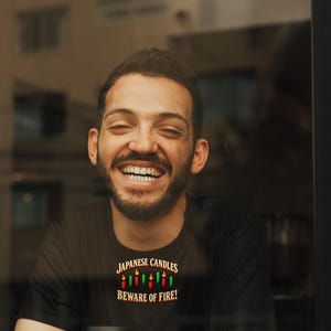 May include: A black t-shirt featuring a graphic of five colourful candles with flames and the text "JAPANESE CANDLES BEWARE OF FIRE!" printed in white and yellow. The shirt is paired with a white disposable coffee cup with a black lid.