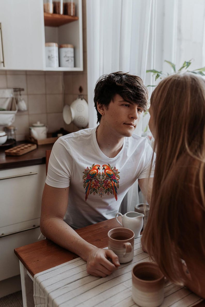 May include: A white short-sleeved t-shirt featuring a colorful graphic print on the chest. The design displays two vibrant parrots facing each other, surrounded by detailed floral and leaf patterns in shades of red, orange, yellow, and green.