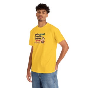 May include: A bright yellow short-sleeve t-shirt featuring a graphic print with the text "you're great from far... far from great." The design includes a small illustration of a person and several small red circular icons below the text.