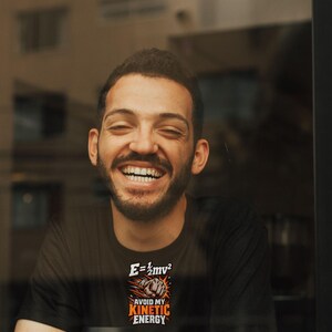 Puede incluir: Una camiseta negra con un estampado gr&aacute;fico que muestra la ecuaci&oacute;n f&iacute;sica E=1/2mv2 sobre un pu&ntilde;o cerrado y el texto AVOID MY KINETIC ENERGY en letras de color naranja y blanco.