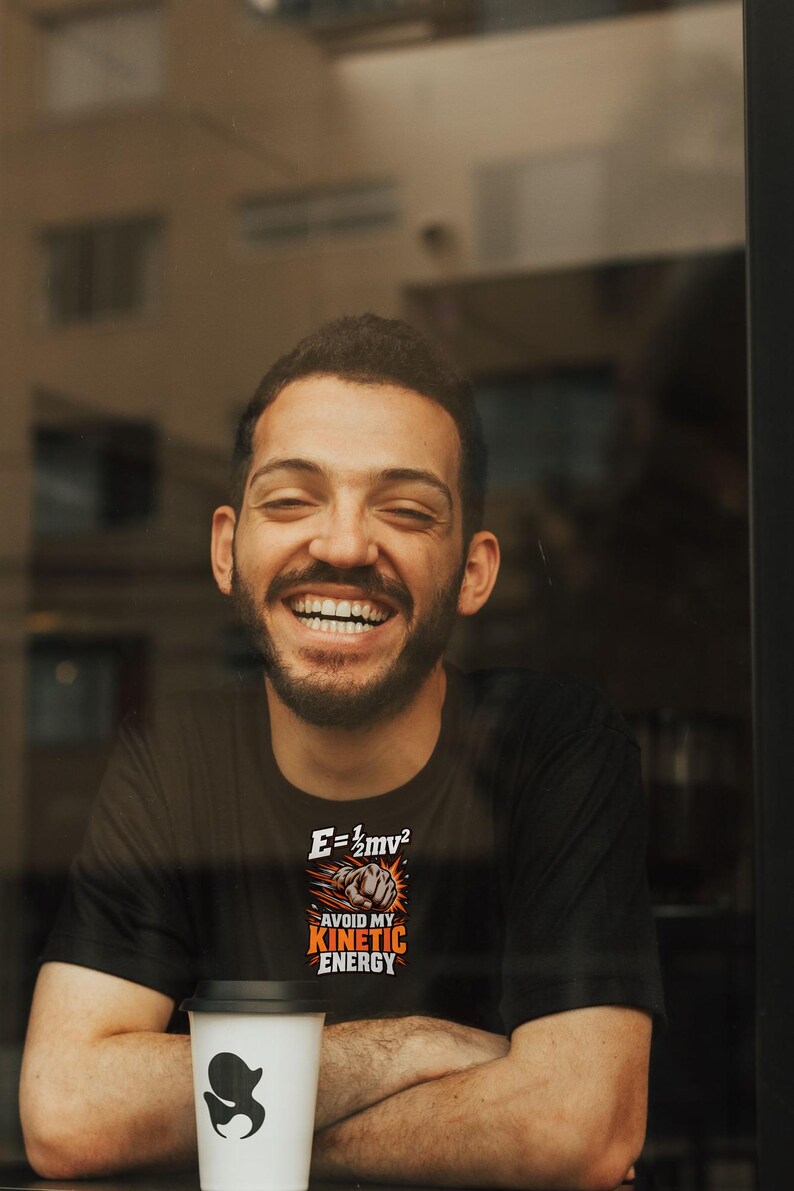 Puede incluir: Una camiseta negra con un estampado gr&aacute;fico que muestra la ecuaci&oacute;n f&iacute;sica E=1/2mv2 sobre un pu&ntilde;o cerrado y el texto AVOID MY KINETIC ENERGY en letras de color naranja y blanco.
